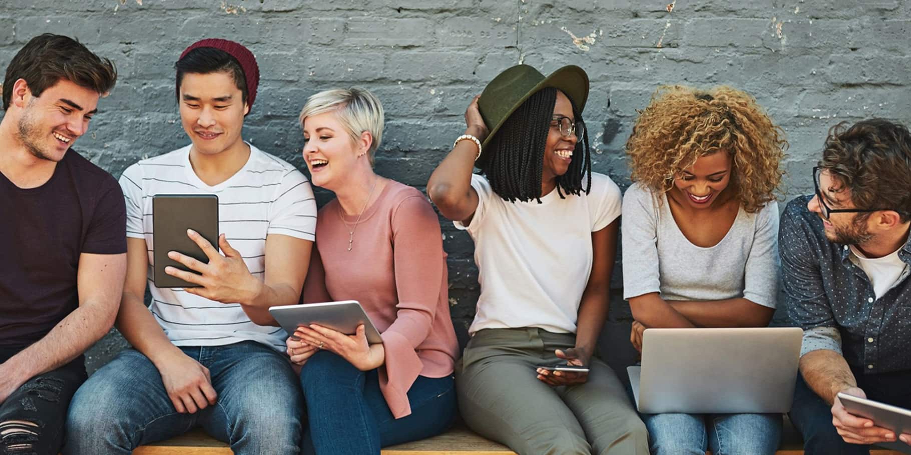 A group of young people sitting against a wall looking at each other's smart devices and laptops.
