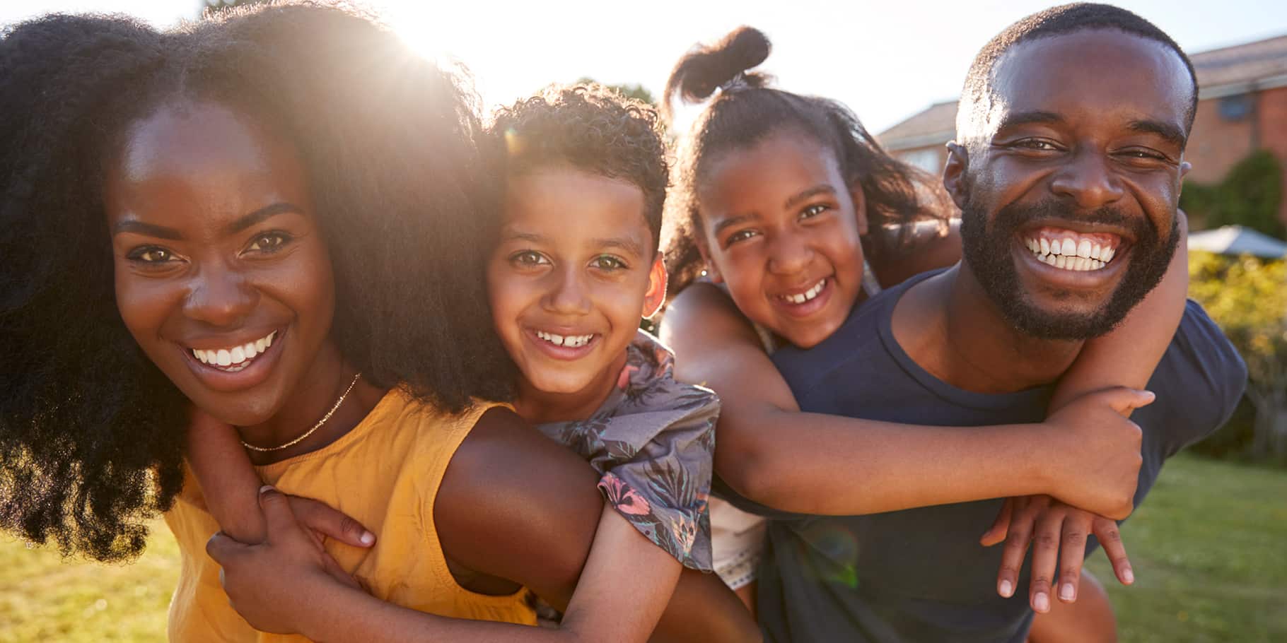 A young smiling family in a grass field with the kids riding the backs of the parents.