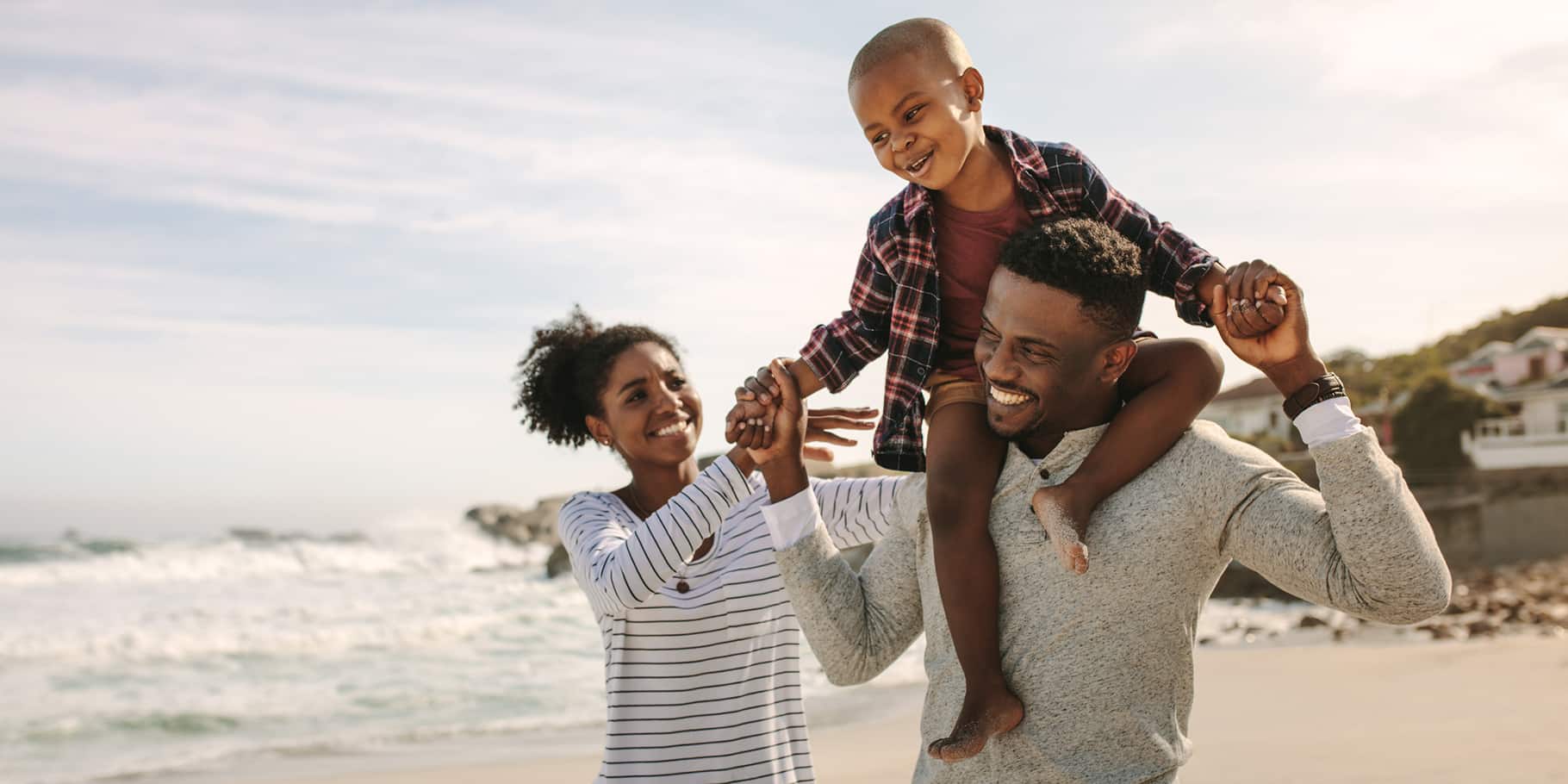 A young family on a beach with the father carrying his son on his shoulders.